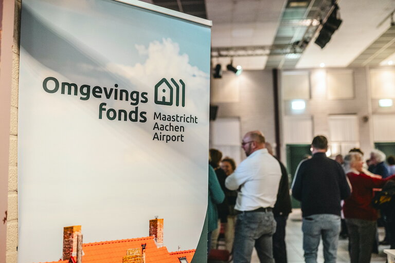 Banner van het Omgevingsfonds MAA met doorkijk naar mensen in de zaal