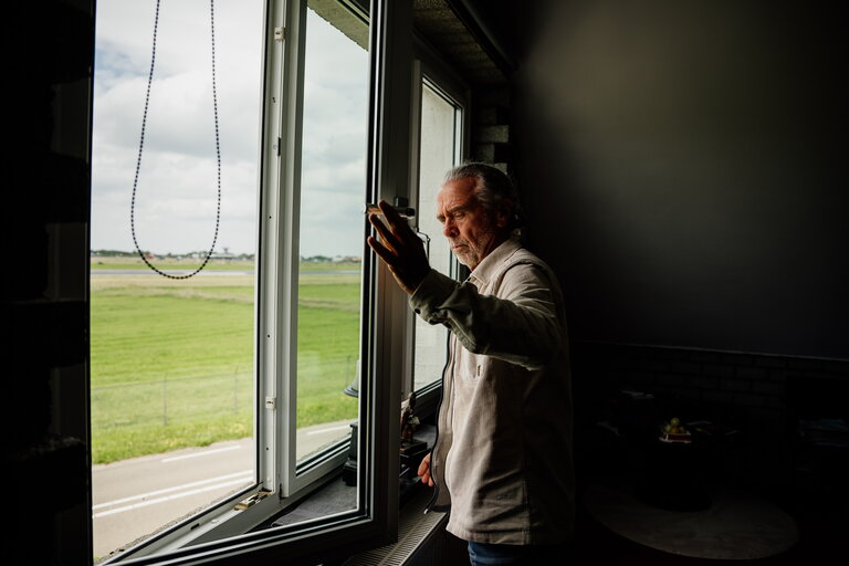 Omwonende John Huizinga voor een raam in zijn woning, door het raam is de luchthaven te zien